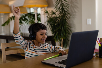 Happy biracial boy using headphones and laptop for online lesson at home, raising hand