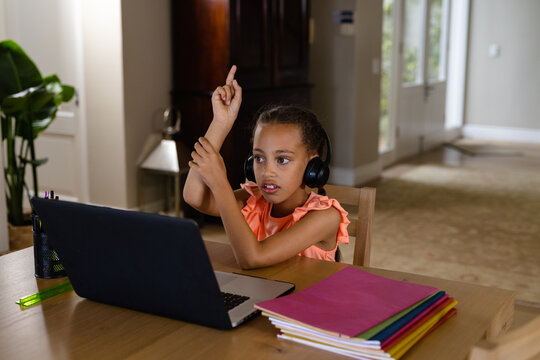 Biracial girl using headphones and laptop for online lesson at home raising hand