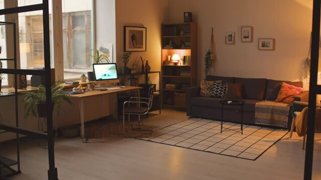 No people shot of cozy living room interior with warm light, pictures on walls, couch and office desk with turned on desktop computer on it