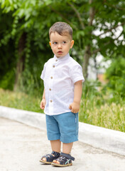 Fototapeta premium adorable little baby boy in fashionable clothes posing outside against wall or green nature trees grass.amazed shocked face.toddler child kid holding hand on building pillar