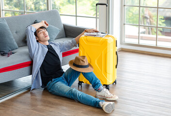 Fototapeta na wymiar Asian cheerful happy male traveler in casual outfit with hat sitting smiling on living room floor holding trolley luggage map passport tickets preparing for summer travel holiday weekend vacation