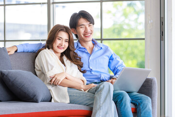 Obraz premium Asian happy cheerful joyful husband and wife lover couple family sitting smiling cuddling together on cozy sofa couch watching streaming movie via laptop notebook computer in living room at home
