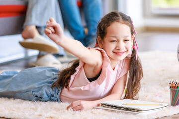 Closeup Asian young little cheerful girl laying down on carpet floor smiling writing painting using color pencils together raised hands with father and mother sitting on sofa blurred background.