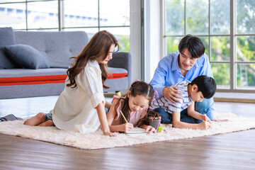 Asian happy cheerful joyful family husband and wife lover couple sitting on carpet floor helping teaching little boy son and girl daughter painting drawing cartoon with color pencils in living room