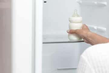 Bottle feeding full of milk in cropped woman hand putting it into refrigerator for storage