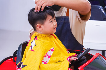 Happy Asian kid boy 2 years old on high chair in toy car shape is looking at tv while having haircut at children barbershop
