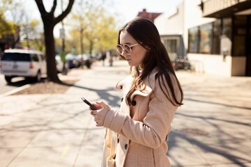 Young female adult stands outside in the autumn, engrossed in her smartphone, texting