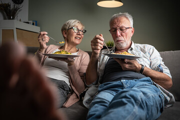 Senior couple sitting on sofa and eating pasta. They're having pleasant conversation and joying in...