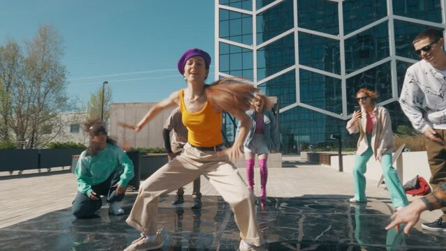 Young And Stylish Female Hip Hop Dancer Freestyling On City Street In the Circle Of Fashionable Friends. Group Of Young People Supporting Professional Newstyle Performer, Filming Her On Smartphone.