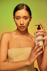product presentation, skin care product, young asian woman with bare shoulders holding cosmetic bottle with body lotion and posing on green background, glowing skin concept