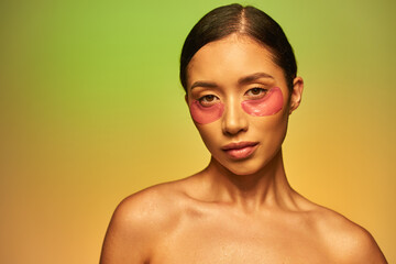 skincare campaign, young asian woman with brunette hair and bare shoulders posing and looking at camera on green background, face care, moisturizing eye patches, glowing skin