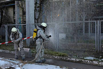 Rescuers in protective rubber suits and masks watering plant territory with syringe. Rescue team training in decontamination