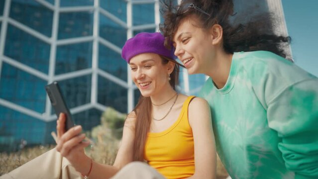 Two Young And Stylish Female Friends Talking On City Street And Using Smartphone To Scroll Social Media Posts. Two Trendy Women Discussing Fashion, News, and Drama Outside In Downtown. Youth Culture.