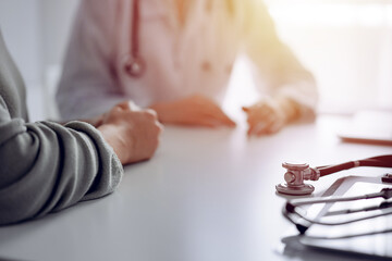 Fototapeta premium Doctor and patient are sitting and discussing something at the desk in the clinic office. The focus is on the stethoscope lying on the table, close up. Perfect medical service and medicine concept
