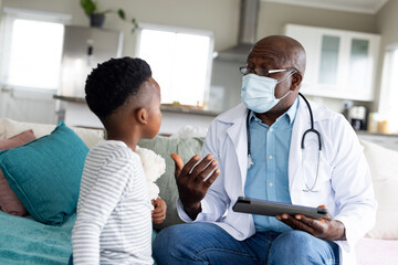 Senior african american male doctor in face mask using tablet talking to boy patient