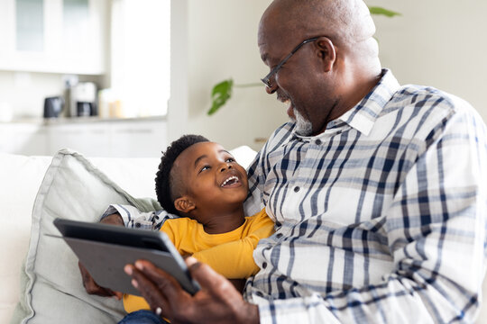 Happy african american grandfather and grandson sitting on sofa and using tablet - Powered by Adobe