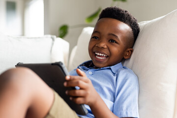 Happy african american boy sitting on sofa and using tablet