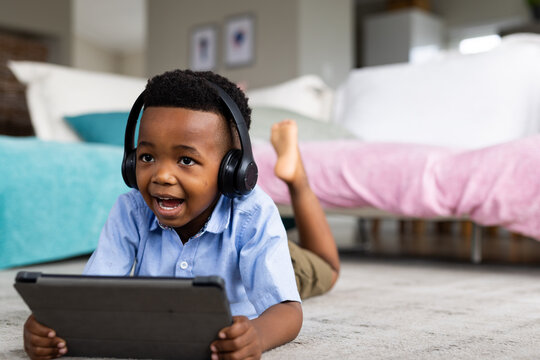 Happy african american boy wearing headphones, lying on floor and using tablet