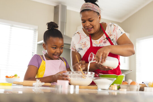 Happy african american mother and daughter baking cupcakes, adding ingredients into bowl in kitchen