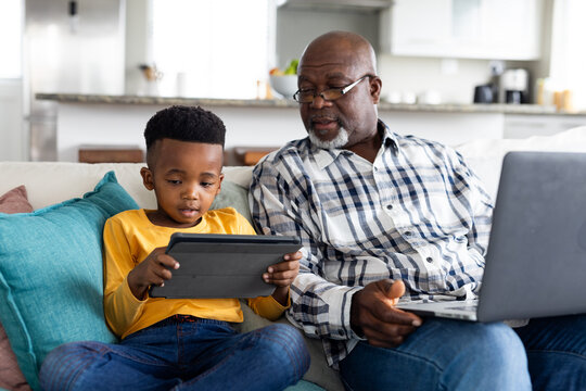 Happy african american grandfather and grandson sitting on sofa, using tablet and laptop - Powered by Adobe