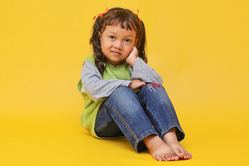 Full body image of cute Asian baby girl sitting with hand on cheek isolated on yellow background