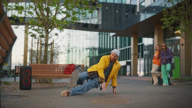 Young Stylish B-boy Breakdancing On City Street Among Modern Buildings In Urban Area. Fashionable Group Of Friends Relaxing On Background, Supporting Breakdance Performer Practising Choreography.