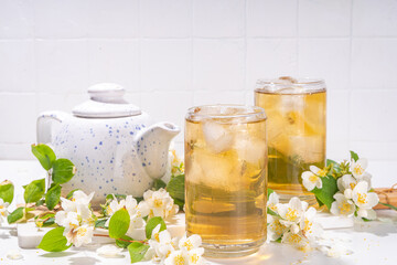 Organic herbal health summer cold drink. Jasmine flowers green tea, in glass, with ice cubes, teapot, on white table, with blooming jasmine flowers 