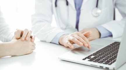 Doctor and patient sitting near each other at the desk in clinic. The focus is on female physician's hands using laptop computer, close up. Medicine concept