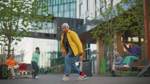 Young Stylish B-boy Breakdancing On City Street Among Modern Buildings In Urban Area. Fashionable Group Of Friends Chilling On Background, Supporting Breakdance Performer Practising Choreography.