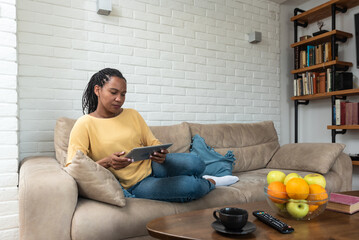 Young African American woman sit relax on couch in living room browsing Internet shopping online on tablet, happy biracial female rest on sofa at home texting messaging on modern pad gadget