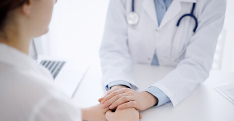 Doctor and patient sitting opposite each other at the table in clinic office. The focus is on female physician's hands reassuring woman, only hands, close up. Medicine concept