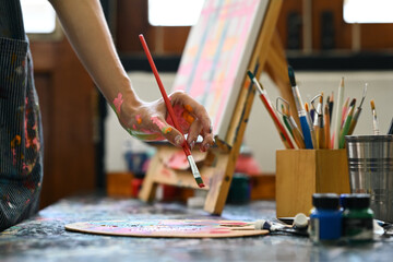 Cropped image of artist standing in front of an easel and dipping brush into color palette