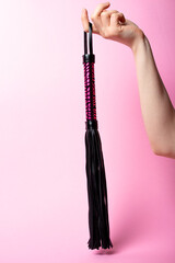 female hand holds a black leather whip with a pink handle on a pink background. Place for text. Sex shop concept. Copy space