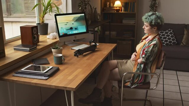 Side view medium shot of Caucasian girl in her early 20s sitting at wooden desk by window with her arm prosthesis lying on it