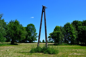 A close up on a power pylon made out of concrete standing in the middle of a dense field, meadow or pastureland, next to some orchard, forest or moor, spotted during summer in Poland