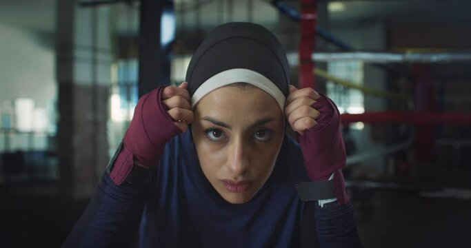 Slow Motion Portrait of Young Determined Arab Woman Adjusting her Hijab in front of the Camera Before Practicing Shadow Boxing in Gym. Strong Muslim Woman Working Out and Exercising Near Boxing Ring