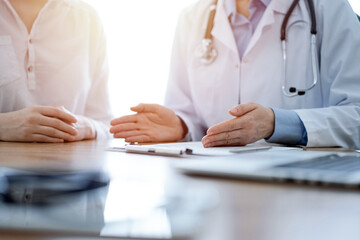 Doctor and patient discussing something while sitting near each other at the wooden desk in clinic, close up of hands. Medicine concept