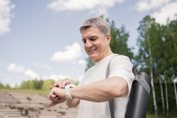 Uses a fitness watch an elderly adult male retired gray-haired workout in the park, doing yoga .