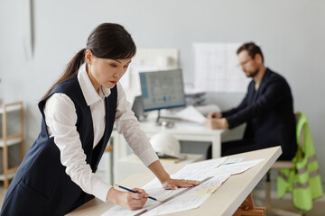 Obraz premium Side view portrait of young woman as engineer drawing blueprints and plans at table in office, copy space