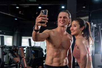 Fototapeta na wymiar Caucasian male trainer holding smartphone taking selfie with Asian woman wearing sports bra as students exercising together in sport club gym