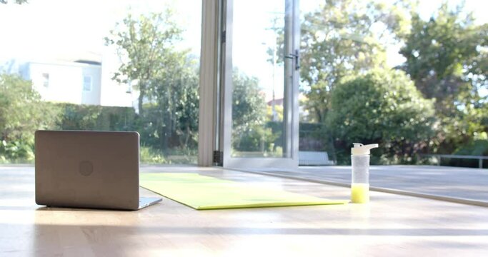 Close up of yoga mat, laptop and bottle with juice on floor at sunny home, slow motion