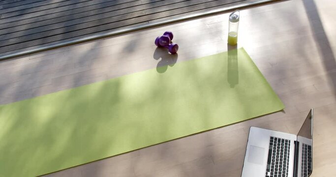 High angle view of yoga mat, laptop, bottle with juice and weights on floor at home, slow motion
