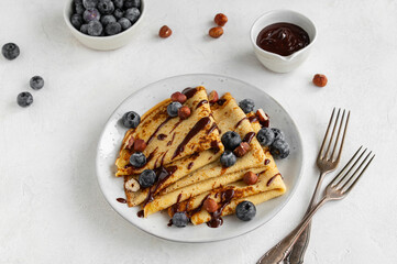 Crepes, blinis or thin pancakes with blueberries, hazelnuts and chocolate sauce on a white plate at white stone table. Maslenitsa traditional festival meal. Delicious breakfast.