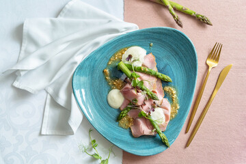 Top view of an appetizer with asparagus, taleggio, peanuts and rosemary ham on a blue plate. White and pink cloth, white napkin and golden cutlery on the table.