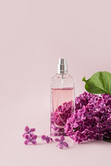 A chic bottle of women's perfume or cosmetic sparea on a pink background with a live branch of purple lilac. vertical view, a copy space.