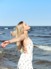 Happy blonde beautiful woman on the ocean beach standing in a white summer dress, open arms - 618478218