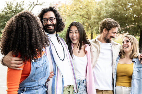Multiethnic group of friends having fun together looking each other in the street.Diverse young women and men hugging each other laughing and carefree staring at each other outside.