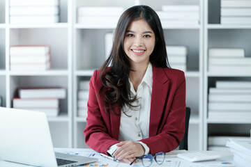 Beautiful asian businesswoman working on laptop and calculating with financial calculator from statistics data graphs, charts. Successful business results in modern office wear red shirt.