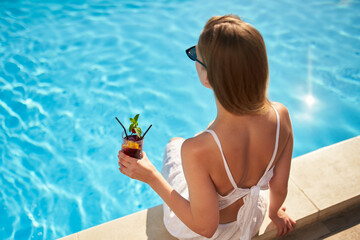Blonde woman drinks refreshing cocktail sunbathing and sitting near swimming pool at tropical spa....