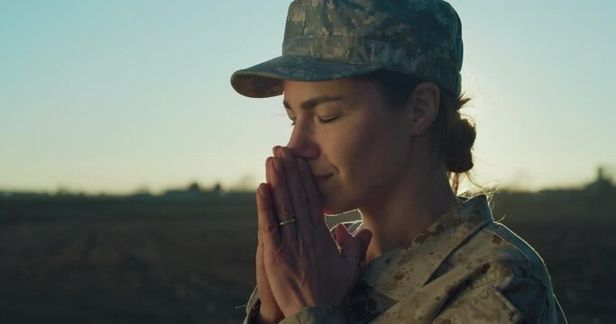 Cinematic Close Up Portrait of Young Female Patriotic Soldier Praying with Hope While Standing in Open Field During Sunset. Woman in Camouflage Military Uniform Hoping for Peace and Freedom  